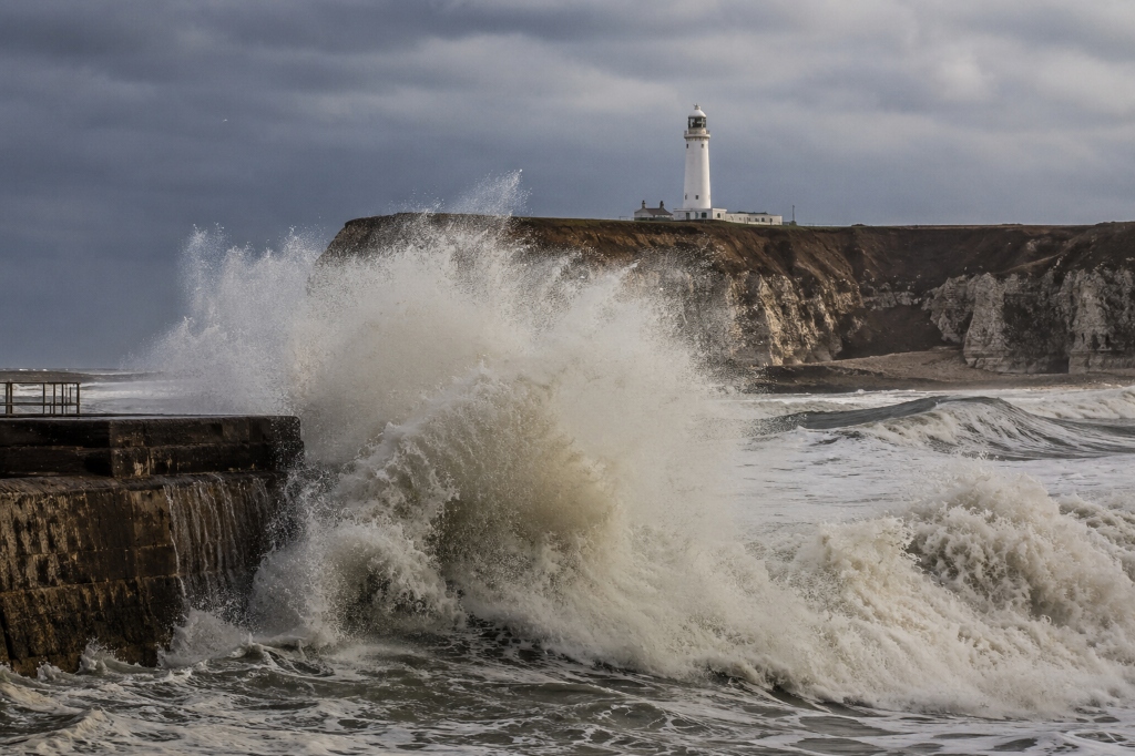 Waves breaking over a harbour wall — Flamborough Head lighthouse on the cliff beyond, North Sea in winter